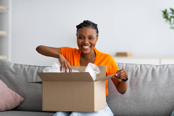 Young Afro woman opening cardboard box, satisfied with good delivery service, receiving online store order at home - Foto, Imagen