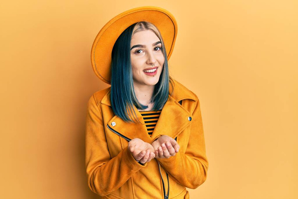 黄色の帽子と革のジャケットを身に着けている若い現代の女の子は一緒に手で笑みを浮かべて手のひらを受信またはジェスチャーを与える。保持し保護する  - 写真・画像