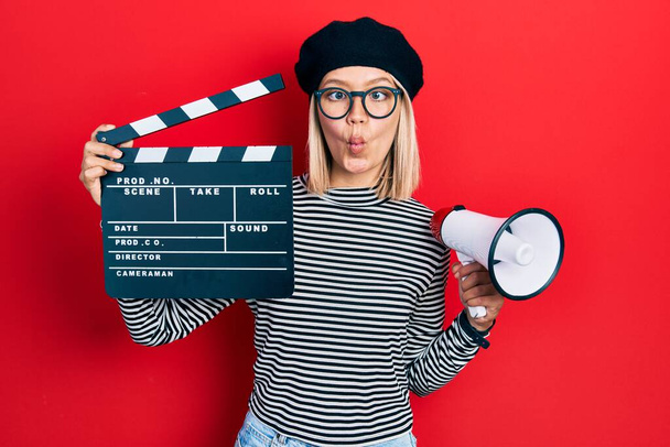 Beautiful blonde woman holding video film clapboard and megaphone making fish face with mouth and squinting eyes, crazy and comical.  - Photo, Image