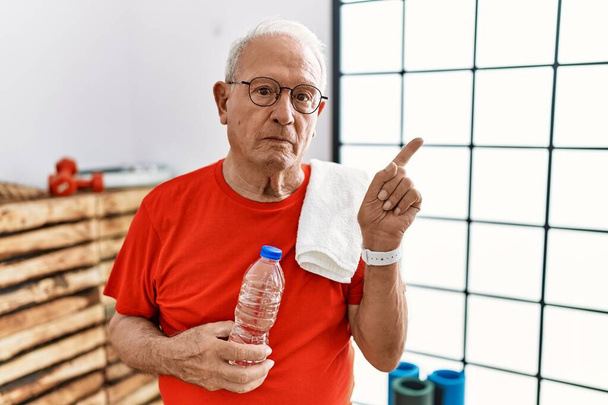 Senior man draagt sportkleding en handdoek op de sportschool wijzen met de hand vinger naar de zijkant tonen reclame, ernstige en rustige gezicht  - Foto, afbeelding