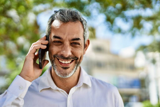Седовласый мужчина средних лет улыбается счастливо разговаривая по смартфону в городе. - Фото, изображение