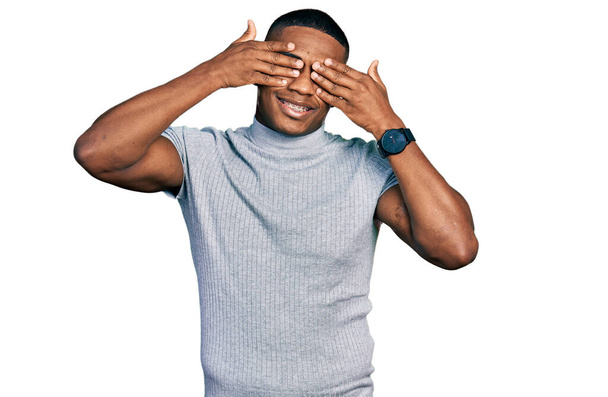 Young black man wearing casual t shirt covering eyes with hands smiling cheerful and funny. blind concept.  - Photo, Image