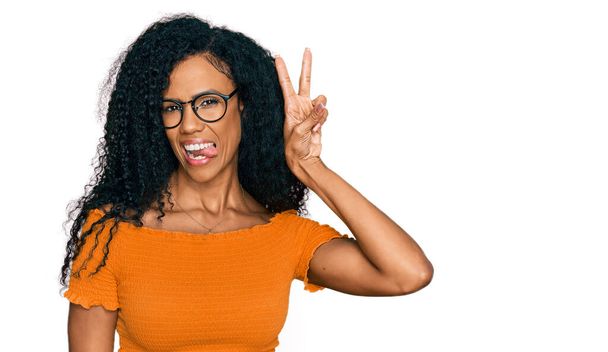 Középkorú afro-amerikai nő alkalmi ruhát és szemüveget visel, mosolyogva, boldog arccal kacsint a kamerára, győzelmi jelet téve. Második..  - Fotó, kép