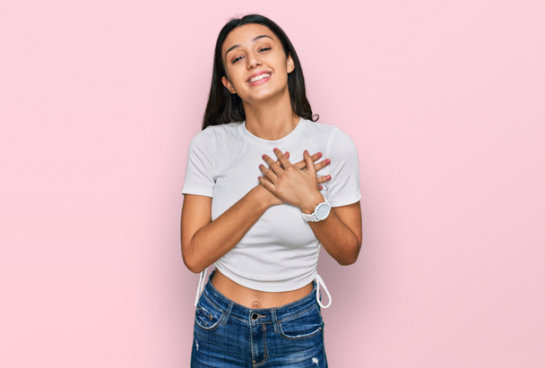 Молодая латиноамериканка в обычной белой футболке улыбается с закрытыми глазами на груди и благодарным жестом на лице. концепция здоровья.  - Фото, изображение