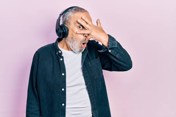 Красивый пожилой мужчина с бородой слушает музыку с помощью наушников, подглядывая в ударные закрывающие лицо и глаза рукой, глядя сквозь пальцы со смущенным выражением лица.  - Фото, изображение