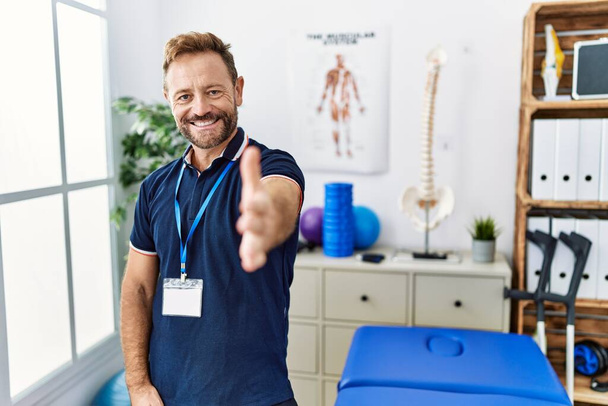 Hombre fisioterapeuta de mediana edad que trabaja en la clínica de recuperación del dolor sonriendo amable ofreciendo apretón de manos como saludo y bienvenida. negocios exitosos.  - Foto, Imagen