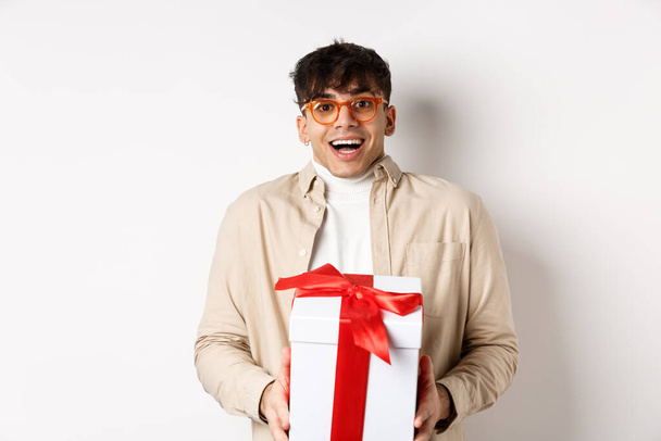 Surprised happy guy receiving a gift, holding present box and smiling amazed, standing on white background - Foto, imagen