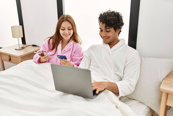 Giovane coppia sorridente felice con laptop, smartphone e carta di credito in camera da letto. - Foto, immagini