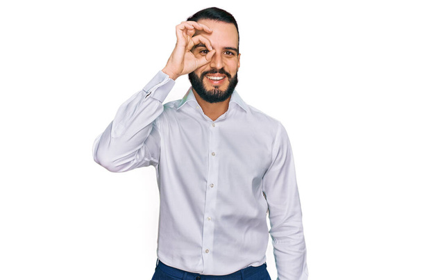 Young man with beard wearing business shirt doing ok gesture with hand smiling, eye looking through fingers with happy face.  - Foto, afbeelding