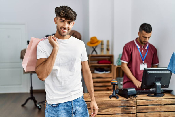 Jonge Latijns-Amerikaanse klant glimlachend gelukkig houden boodschappentas in kledingwinkel. - Foto, afbeelding