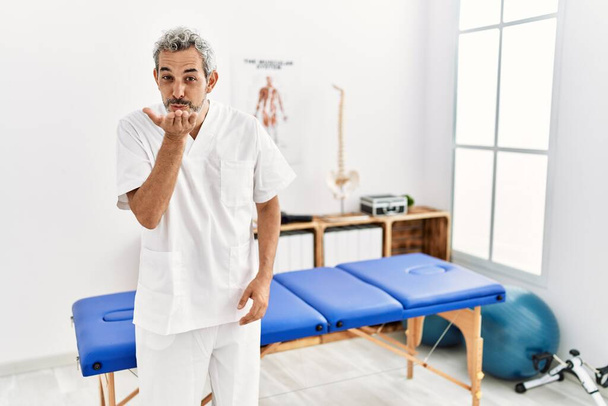 Middle age hispanic therapist man working at pain recovery clinic looking at the camera blowing a kiss with hand on air being lovely and sexy. love expression.  - 写真・画像