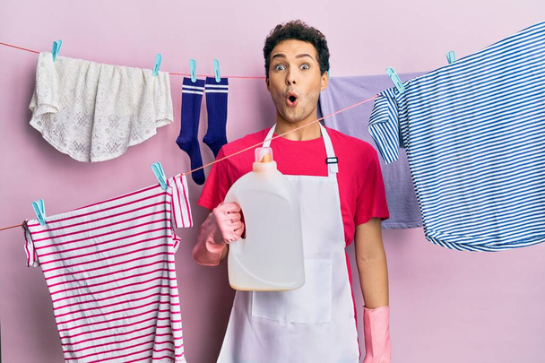 Hombre hispano guapo lavando la ropa sosteniendo botella de detergente asustado y sorprendido con la boca abierta para sorpresa, cara de incredulidad  - Foto, Imagen