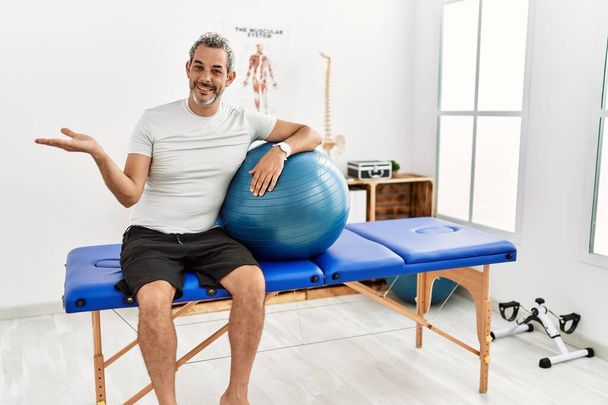 Middle age hispanic man at pain recovery clinic holding pilates ball smiling cheerful presenting and pointing with palm of hand looking at the camera.  - Fotoğraf, Görsel