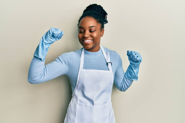 Mujer afroamericana con el pelo trenzado usando delantal más limpio y guantes que muestran los músculos de los brazos sonriendo orgulloso. concepto de fitness.  - Foto, imagen