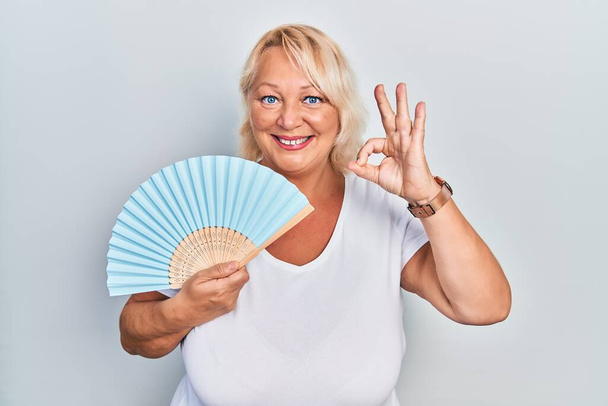 Mujer rubia de mediana edad agitando ventilador de mano enfriamiento de aire en verano haciendo signo ok con los dedos, sonriendo gesto amistoso excelente símbolo  - Foto, Imagen