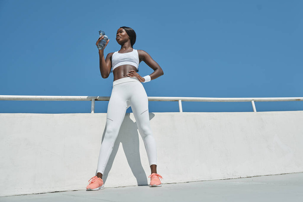 Low angle view of beautiful young African woman in sports clothing drinking water while standing outdoors - Photo, Image