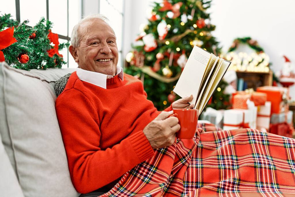 Senior grey-haired man drinking coffee and reading book celebrating christmas at home. - Photo, Image