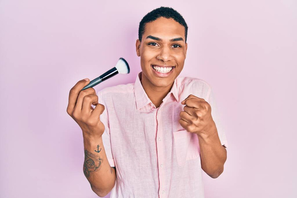 Young african american guy holding makeup brush screaming proud, celebrating victory and success very excited with raised arm  - Photo, Image