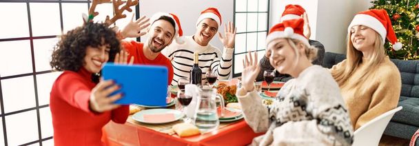 Grupa młodych ludzi przy świątecznej kolacji i rozmowie wideo za pomocą touchpada w domu. - Zdjęcie, obraz