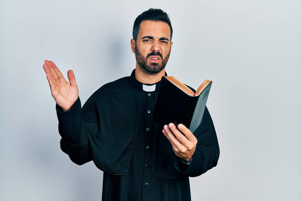 Handsome hispanic priest man with beard holding bible and christian cross clueless and confused expression. doubt concept.  - Photo, Image