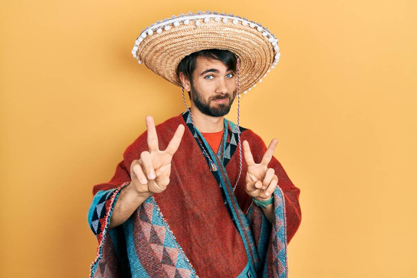 Jeune homme hispanique tenant un chapeau mexicain souriant regardant la caméra montrant des doigts faisant signe de victoire. numéro deux.  - Photo, image