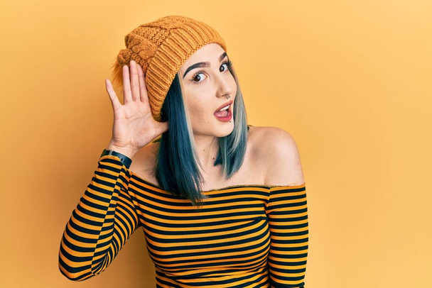 Νεαρή μοντέρνα κοπέλα που φοράει μάλλινο καπέλο χαμογελώντας με το χέρι πάνω από το αυτί ακούγοντας μια ακρόαση για φήμες ή κουτσομπολιά. έννοια της κώφωσης.  - Φωτογραφία, εικόνα