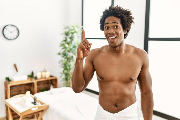 Jeune homme afro-américain torse nu portant une serviette debout au centre de beauté pointant du doigt avec une idée réussie. Sorti et heureux. numéro un.  - Photo, image