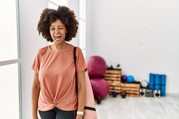 African american woman with afro hair holding yoga mat at pilates room sticking tongue out happy with funny expression. emotion concept.  - Фото, изображение