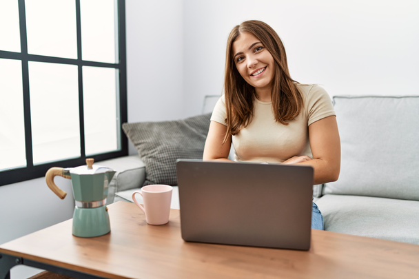自宅でノートパソコンを使用して若いブルネットの女性は、カメラを見ている交差腕で笑顔のコーヒー幸せな顔のカップを飲んでいます。正の人.  - 写真・画像