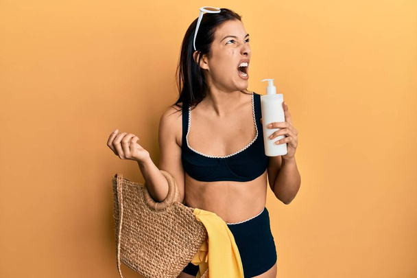 Young latin woman wearing bikini and hat holding summer wicker handbag and sunscreen angry and mad screaming frustrated and furious, shouting with anger. rage and aggressive concept.  - Foto, Imagem