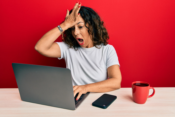 Belle femme d'âge moyen travaillant au bureau à l'aide d'un ordinateur portable stressé et frustré avec la main sur la tête, visage surpris et en colère  - Photo, image
