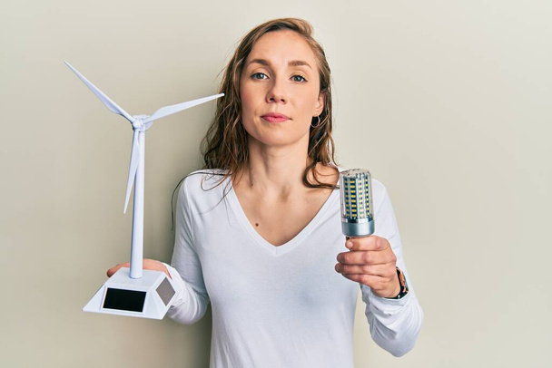 再生可能エネルギー用のソーラー風車を持つ若いブロンドの女性と顔に深刻な表情でリラックス電球を導きました。カメラを見ているだけで.  - 写真・画像