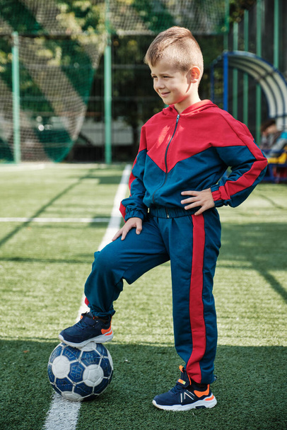Een kleine jongen zette zijn voet op de bal op het voetbalveld. Kleine jongen in kleurrijke trainingspak. Herfst lente sport kinderen mode. - Foto, afbeelding