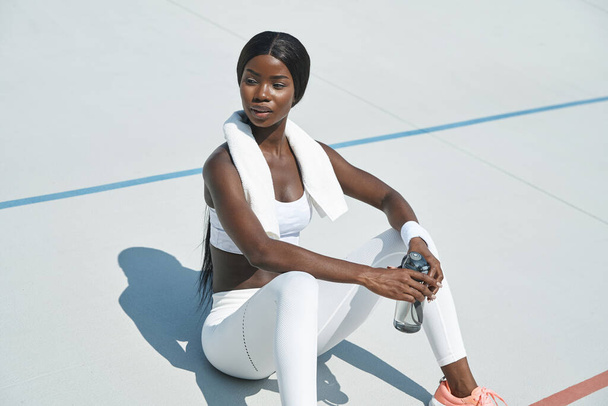 Beautiful young African woman in sports clothing holding bottle with water while sitting on sports track outdoors - Φωτογραφία, εικόνα