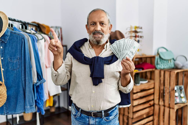 Beau senior au magasin de détail tenant des billets en dollars souriant heureux pointant avec la main et le doigt sur le côté  - Photo, image