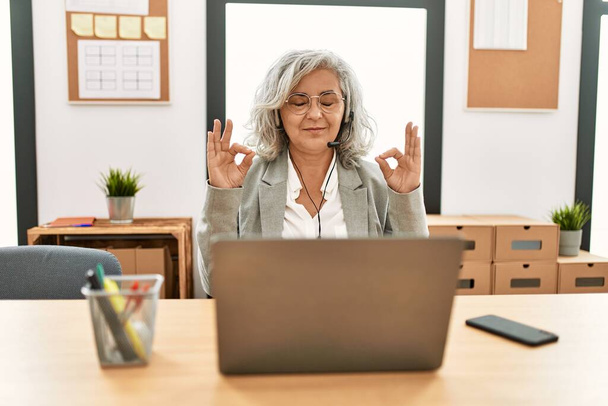 Бізнес-леді середнього віку, яка сидить на столі, працюючи з ноутбуком в офісі, розслабляється і посміхається очима, закритими, роблячи медитаційний жест пальцями. йога концепція
.  - Фото, зображення
