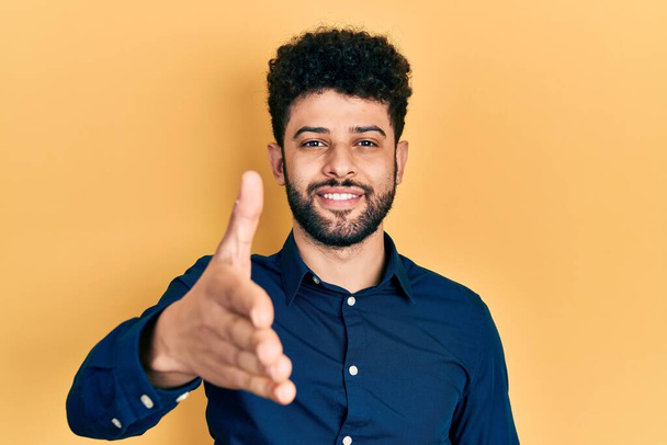 Junger arabischer Mann mit Bart trägt lässiges Hemd und lächelt freundlich und reicht zur Begrüßung Handschlag. Erfolgreiches Geschäft.  - Foto, Bild