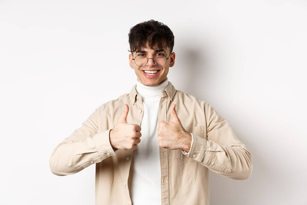 Vrolijke hipster man in bril tonen duimen omhoog en glimlachen, als goed product, aanbevelen of lof reclame, staande op witte achtergrond - Foto, afbeelding