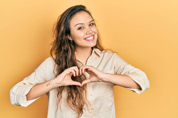 Young hispanic girl wearing casual clothes smiling in love doing heart symbol shape with hands. romantic concept.  - Photo, image