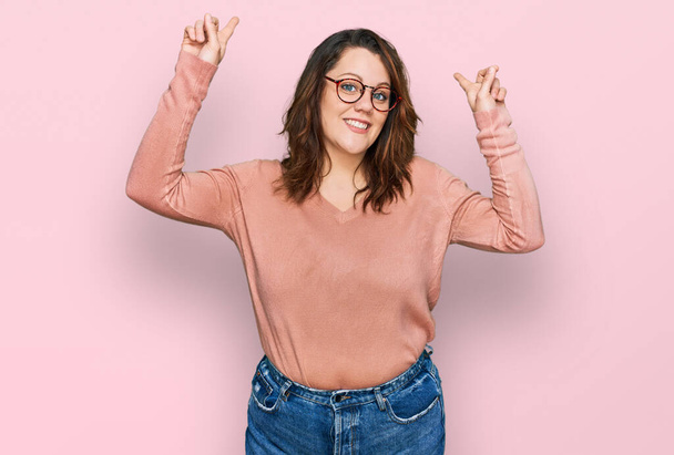 Young plus size woman wearing casual clothes and glasses smiling amazed and surprised and pointing up with fingers and raised arms.  - Foto, afbeelding