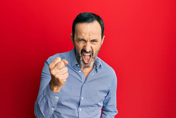 Orta yaşlı İspanyol bir adam, günlük kıyafetler giyiyor kızgın ve kızgın. Öfkeyle bağırırken yumruğunu kaldırıyor. öfke ve agresif konsept.  - Fotoğraf, Görsel