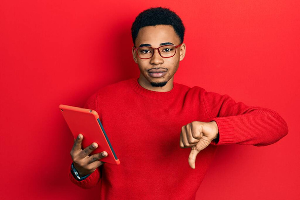 Young african american man using touchpad device with angry face, negative sign showing dislike with thumbs down, rejection concept  - 写真・画像