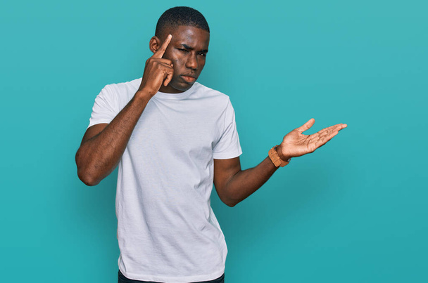 カジュアルな白いTシャツを着た若いアフリカ系アメリカ人の男性は、コピースペースと額に指を指すことを示す開いてヤシの木に混乱し、悩まされました。考えてみてください.  - 写真・画像