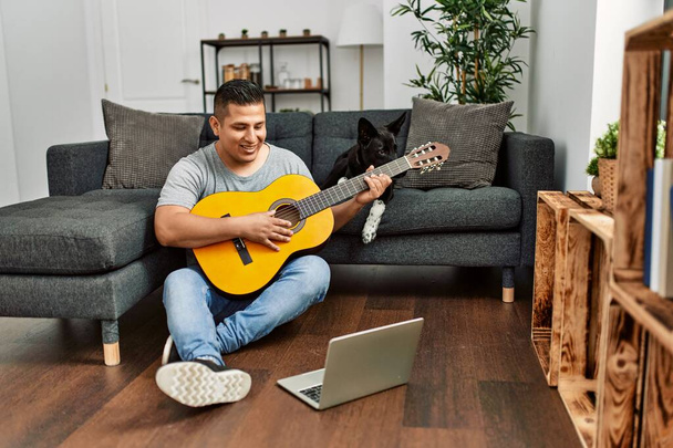 Νεαρός Ισπανόφωνος έχοντας onlinr μάθημα κλασικής κιθάρας κάθεται στο πάτωμα με το σκυλί στο σπίτι. - Φωτογραφία, εικόνα