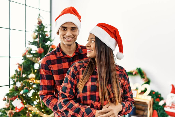 jong latijn paar glimlachen gelukkig en knuffelen stand-by kerstboom thuis. - Foto, afbeelding
