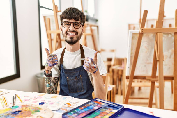 Hispanischer Mann mit Bart auf dem Erfolgsschild des Kunststudios macht eine positive Geste mit der Hand, Daumen hoch lächelnd und glücklich. Fröhlicher Ausdruck und Siegergeste.  - Foto, Bild