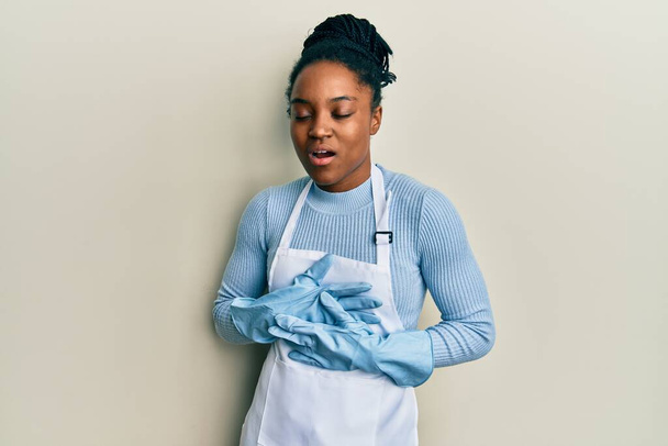 African american woman with braided hair wearing cleaner apron and gloves with hand on stomach because nausea, painful disease feeling unwell. ache concept.  - Фото, зображення