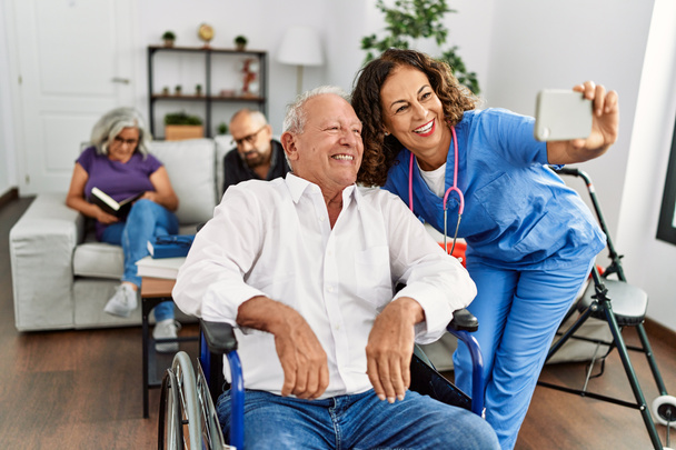 Médico de mediana edad mujer y hombre jubilado sentado en silla de ruedas hacer selfie por el teléfono inteligente en el hogar de ancianos. - Foto, imagen