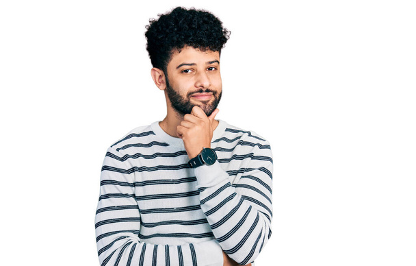 Hombre árabe joven con barba llevando suéter de rayas casual mirando confiado a la cámara sonriendo con los brazos cruzados y la mano levantada en la barbilla. pensar positivamente.  - Foto, Imagen