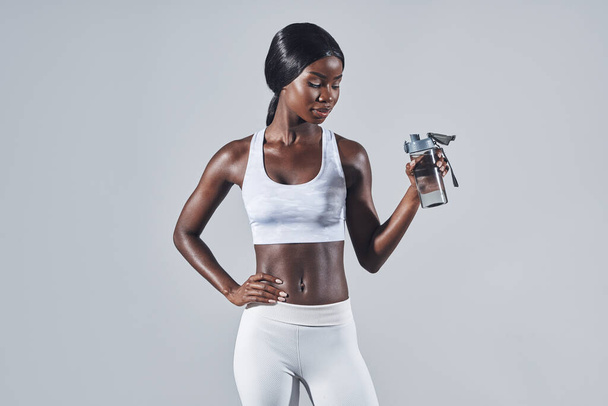 Beautiful young African woman in sports clothing drinking water while standing against gray background - Φωτογραφία, εικόνα